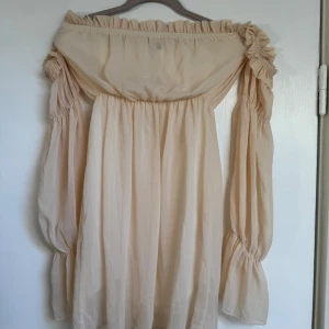 Beige offshoulder klänning med volang - Säljer en superfin beige offshoulder klänning med långa puffiga ärmar och volangdetaljer vid axlar och ärmslut. Klänningen har en luftig och flowy passform, perfekt för varma dagar. Materialet känns lätt och skönt mot huden.