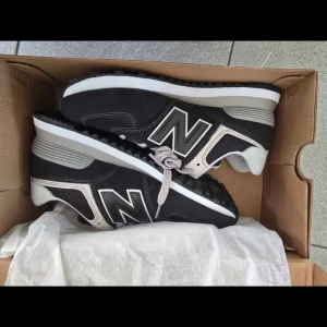 New Balance 574 svarta sneakers - Svarta New Balance 574 sneakers med grå och vita detaljer. Klassisk retrostil med stor N-logga på sidan, snörning och platt sula. Ovandel i textil och mocka för en sportig vibe. Perfekta för dig som gillar streetwear och bekväma skor.