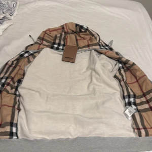 Grå zip-hoodie från Burberry - Grå hoodiejacka från Burberry med dragkedja, klassisk rutig huva i beige, svart, rött och vitt. Broderad Burberry-logga på bröstet och fickor framtill. Mjuk bomullskänsla och snygga detaljer som snörning i huvan.