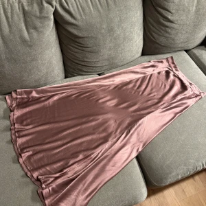 Rosa satin långkjol från Zara - Säljer en lång, glansig satinkjol från Zara i en snygg lila-rosa nyans. Kjolen har en mjuk, följsam passform och är perfekt för dig som gillar en stilren och trendig look. Materialet är lent och skimrande, vilket ger kjolen en lyxig känsla. Det finns ett litet fel (syns på bild 3), men det är knappt märkbart.