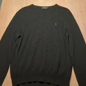 Svart tröja från Polo Ralph Lauren - Svart långärmad tröja från Polo Ralph Lauren med klassisk rund hals och lila broderad logga på bröstet. Tröjan är i mjuk ull och har en normal passform, perfekt till jeans eller chinos. Enkel och stilren design som funkar året runt.