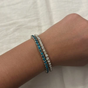 Strassarmband med blå och vita stenar - Två glittriga armband med runda strasstenar, ett med klara vita stenar och ett med intensivt blå stenar. Båda har silverfärgad metall och elastisk design som gör dem enkla att ta på. Perfekt för dig som gillar att sticka ut med lite extra bling.