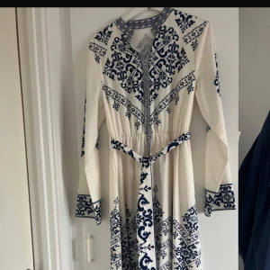 Vit och blå långklänning med mönster - Långklänning i vit med marinblått mönster och knytband i midjan. Klänningen har lång ärm, rundad halsringning och detaljerade blå mönster längs ärmslut, midja och nederkant. Perfekt för dig som gillar bohemisk och stilren look. Och perfekt för hijabi kvinnor. Den är helt ny