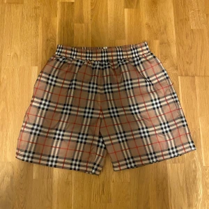 Rutig Burberry badshorts i beige och rött - Snygga shorts från Burberry i klassiskt rutigt mönster med beige, svart, vitt och röda detaljer. De har elastisk midja och är tillverkade i bomull med meshfoder. Perfekta för sommaren och ger en clean och trendig look.