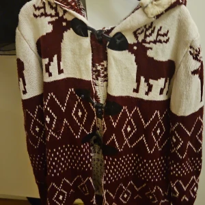 Vinröd och beige stickad kofta med huva - Mysig stickad kofta i vinrött och beige med huva och pälsimitation. Koftan har ett klassiskt vintermönster med renar och geometriska former, samt snygga svarta knäppningar framtill. Perfekt för kalla dagar när du vill ha något både varmt och snyggt.