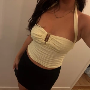 Ljusgul ribbad halterneck topp - Supersöt ljusgul ribbad halterneck topp med knyt i nacken och snygg detalj framtill i form av en plastspänne. Toppen är croppad och har en smickrande passform som framhäver axlarna. Perfekt för varma dagar och somriga vibes. Aldrig använd