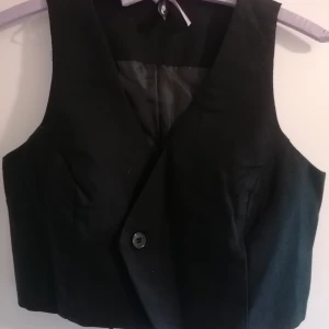 Svart kostymväst  - Stilren svart väst från FB Sister i kostymstil med knäppning framtill och justerbart bälte bak. Västen har en klassisk v-ringad form och är fodrad för extra komfort. Perfekt att styla över en skjorta eller t-shirt för en trendig look.                                                         Den är helt ny och oanvänd! Utan tag. 