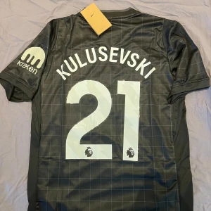 Tottenham Kulusevski 21 tröja - Snygg svart Tottenham Hotspur fotbollströja från Nike med Kulusevski och nummer 21 på ryggen. Tröjan har korta ärmar, diskret rutmönster, AIA som sponsor på bröstet och klubbmärke på vänster sida. Tillverkad i lätt och ventilerande material.
