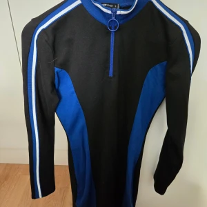 Sportig svart och blå klänning - Cool kortklänning i svart och blått med vita ränder längs ärmarna. Klänningen har lång ärm, ståkrage och en snygg ringformad dragkedja framtill. Materialet är mjukt och stretchigt.