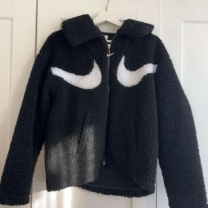 Svart teddyjacka från Nike XS - Svart teddyjacka från Nike i storlek XS med stora vita swoosh-loggor på bröstet. Jackan har dragkedja framtill, klassisk krage och är tillverkad i mjukt fleece-material. Perfekt för dig som vill ha en sportig och trendig look.