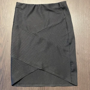 Svart ribbad kortkjol med omlott - Snygg svart kortkjol med ribbad struktur och omlott-detalj framtill. Kjolen har en tight passform och är perfekt för dig som gillar stilrena plagg med en twist. Enkel att matcha med både t-shirt och crop top.