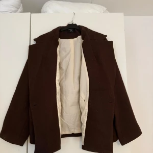 Brun kappa från Lindex XS - Säljer en stilren brun kappa från Lindex i storlek XS. Jackan har klassisk krage, två fickor fram och stängs med en knapp. Insidan är ljus och mjuk, perfekt för höst och vinter. Snygg och enkel att matcha med olika outfits.