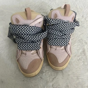 Lanvin sneakers i ljusrosa med mönstrade snören - Lanvin sneakers i ljusrosa skinn med beige detaljer och chunky sula. Skorna har unika breda snören i svartvitt sicksackmönster som verkligen sticker ut. Perforerad tå och hög modell för en cool streetstyle-vibe.