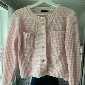 Rosa rutig kofta - Koftan är stickad med rosa och vita rutor, har två bröstfickor och guldknappar framtill. Använd fåtal gånger 