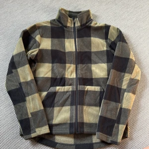 Rutig fleecejacka från Firefly - Mysig fleecejacka från Firefly i svart och grön/beige rutigt mönster. Jackan har hög krage, hel dragkedja framtill och två fickor. Perfekt för kyliga dagar och enkel att slänga på sig över en hoodie eller t-shirt.