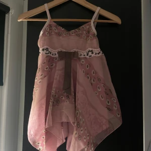 Rosa topp med rosett - Supersöt rosa topp med tunna axelband, omlottkänsla och flowig asymmetrisk nederdel. Toppen har ett unikt mönster, spetsdetalj med paljetter och en stor brun rosett framtill. Perfekt för dig som gillar romantiska och lekfulla plagg. Står 134/140 men passar ocksåXS/S