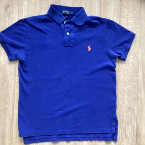 Blå pikétröja från Polo Ralph Lauren - Snygg blå pikétröja från Polo Ralph Lauren med klassisk krage, två knappar och orange broderad logga på bröstet. Modellen är custom fit och har korta ärmar. Perfekt för en stilren och sportig look.Jag kan lägra priset om man vill få för bättre pris