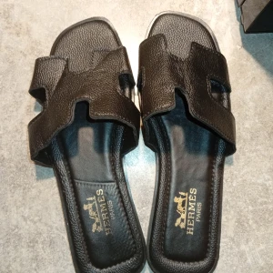 Svarta sandaler  - Svarta sandaler med ikonisk H-formad rem i präglat skinn. Snygg, stilren design med öppen tå och platt sula. Guldtryck Paris-logga på innersulan och Eiffeltornet på yttersulan. Perfekta för en trendig och lyxig look.