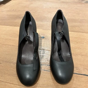 Svarta Mary Jane-klackar Laura Clement - Stilrena svarta Mary Jane-klackar från Laura Clement i skinn med rund tå och justerbart spänne över vristen. Skorna har en stadig hög klack och klassisk siluett som ger en tidlös look. Perfekta för dig som gillar en elegant och enkel stil.
