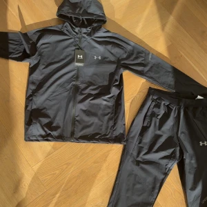  Under Armour - Sportig svart väst från Under Armour med huva och dragkedja framtill. Västen har en stilren design med logga på bröstet och är tillverkad i lätt syntetmaterial som andas. Perfekt för träning och aktiva dagar.