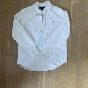 Vit skjorta från Ralph Lauren - Klassisk vit skjorta från Ralph Lauren med broderad logga på bröstet. Skjortan har lång ärm, knappar framtill och är tillverkad i bomull. Snygg och tidlös modell med custom fit som passar till många olika stilar.