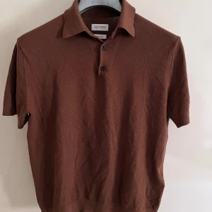 Brun pikétröja från These Glory Days - Säljer en brun pikétröja från These Glory Days i silkeblandning. Tröjan har klassisk krage, korta ärmar och två knappar framtill. Materialet är mjukt och har en diskret struktur som ger en snygg look.