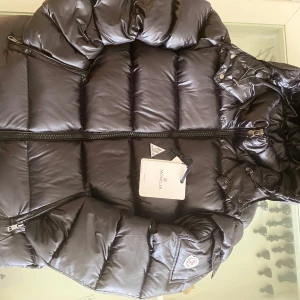 Svart Moncler dunjacka med huva - Säljer en svart Moncler dunjacka med huva och dragkedja. Jackan har klassisk quiltad design, Moncler-logga på ärmen och coolt serietidningsfoder på insidan. Flera fickor med dragkedja och snygga detaljer. Perfekt för kalla dagar och riktigt stilren look. Det är storlek M/S. Jackan passar både Small och Medium
