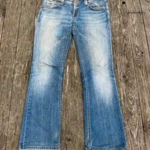 Blå bootcut jeans från Nudie Jeans - Snygga blå bootcut jeans från Nudie Jeans med klassisk femficksdesign och orangea sömmar. Jeansen har en ljus tvätt med slitningar framtill och broderade bakfickor. Tillverkade i mjuk bomullsdenim och passar perfekt för en avslappnad stil. tydligen en unisex modell men skulle ändå säga att de lutet mer åt dam hållet. 