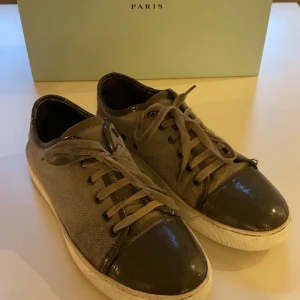 Gråa captoe skor från Lanvin Paris  - Säljer ett par grå skor från Lanvin Paris med snygg mocka på sidorna och glansig tå i mörkgrått. Skorna har snörning och vit sula, perfekt för dig som gillar stilrena och exklusiva sneakers med en twist. Storlek 43/44. Boxen medföljer ej.
