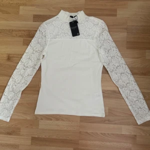 Ny. Vit spetsblus från The Black Label - Supersnygg vit blus från The Black Label med långärmade spetsärmar och spetsdetaljer upptill. Hög hals och figurnära passform. Tillverkad i bomull, polyester och elastan för en bekväm känsla. Perfekt för dig som vill ha något stilrent men ändå unikt.