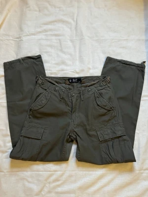 Olivgrön / mörkgröna cargobyxor från Bandit - 👖Produktbeskrivning 👖Snygga mörkgröna cargobyxor från Brandit  👖Köpta från Zalando — varumärket Brandit 👖Nypris är 659kr på Zalando!  👖Midjemått 82cm (41cm x 2). 👖Längd: 106cm 👖Storlek EU30 i jeans. 👖Material: 100% bomull. 👖Med flera stora fickor på sidorna och bak: säkerhetsficka, cargofickor, fickor med lock. 👖Byxorna har raka ben, justerbara band i midjan och vid benslut.  👖Bara lite slitet vid slutet av benen.  Mer information ℹ️ 🧍🏻‍♀️För referens är jag 163cm och tar på mig S/M i storlek.