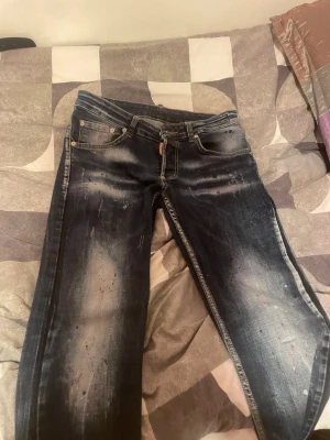 Mörkblå Dsquared2 jeans med slitningar - Säljer ett par mörkblå jeans från Dsquared2 med coola slitningar och färgstänk för en edgy look. Klassisk femficksmodell, knappgylf och tydlig Dsquared2-logga på bakfickan. Jeansen har kontrastsömmar och en straight passform storlek 32 passar mig som 1,80 cm och 70kg