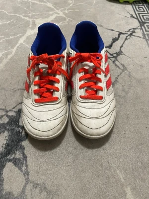 Vita Adidas fotbollsskor med röda  detaljer - Säljer ett par vita Adidas fotbollsskor med klassiska röda ränder och röda skosnören. Insidan är blå och hälen har blå och röda partier. Skorna har rund tå och platt sula, perfekta för dig som vill sticka ut på planen.