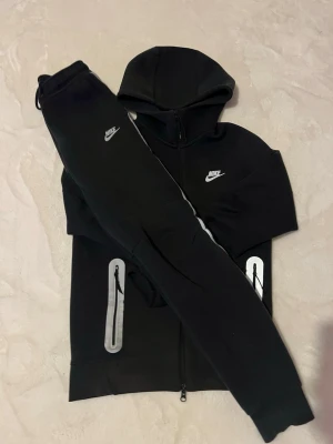 Nike tracksuit - Den har reflexer både fram och bak på hoodien och på byxorna har den reflexer fram och på sidorna. På byxorna finns det en stor ficka med dragkedja på en höger sidan och på vänster en normal ficka. 