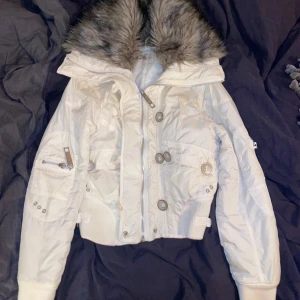 Vit jacka med pälsdetaljer - Säljer en snygg vit jacka från R.B. I i storlek 8 Jackan har en fluffig grå pälsdetalj runt kragen. Den har långa ärmar och flera knappar framtill, samt praktiska fickor med dragkedja och knappar. Jackan är i bra skick. En del saknas på en dragkedja.