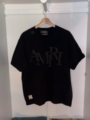 Svart AMIRI t-shirt - Svart t-shirt från AMIRI med stor ton-i-ton logga framtill och en vit AMIRI-detalj nere vid fållen. Klassisk rund hals och korta ärmar. Skön passform och tillverkad i mjuk bomull, perfekt för en clean och stilren look.