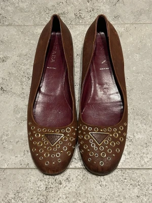 Bruna ballerinaskor från Prada i skinn - Snygga bruna ballerinaskor från Prada i äkta skinn med guldfärgade nitar och ikonisk Prada-triangel framtill. Skorna har rund tå och en klassisk, platt sula. Insidan är vinröd och de är tillverkade i Italien. Storlek 41 men skulle rekommendera för de som har storlek 42 om man inte vill ha strumpor i. Fint bevaren på ovansidan men mer sliten på undersidan