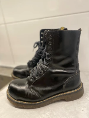 Svarta Dr. Martens boots i skinn - Klassiska svarta Dr. Martens boots i blankt skinn med högt skaft och grov sula. Skorna har snörning hela vägen upp och den ikoniska gula sömmen runt sulan. Tillverkade i England och har AirWair-hälflik med gul text och stålhättar. Skon är i väldigt fint skick bara att innesulan är lite sliten men kan ersättas med en vanlig sula från en annan sko för bästa komfort men funkar utmärkt även utan. Ute sulan är i fint skick utan hål alltså läcker inte något vatten in💕 Hör av er för prisförslag💕💕