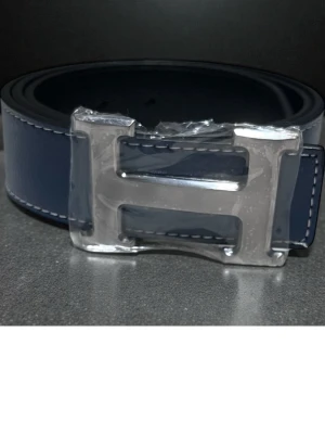 Mörkblå läderbälte med silverdetalj - Snyggt mörkblått bälte i läder med vit kontrastsöm och stor silvrig H-formad metallspänne. Perfekt accessoar för att lyfta din outfit med en clean och modern look.
