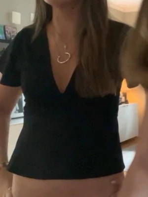 Svart omlottopp med knytband - Svart omlottopp från Gina i mjuk bomull med v-ringning och korta, lite fladdriga ärmar. Toppen har knytband i midjan som ger en snygg passform och en enkel, stilren look. Perfekt att styla med jeans eller kjol för en trendig vibe.