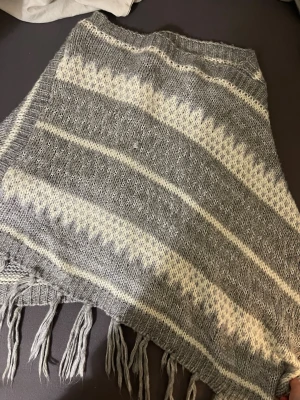 Gråvit stickad poncho med fransar - Mysig stickad poncho i grått och vitt med breda ränder och grafiskt mönster. Ponchon har ribbad kant upptill och dekorativa fransar längs nederkanten. Perfekt för kyliga dagar och enkel att slänga över vilken outfit som helst.
