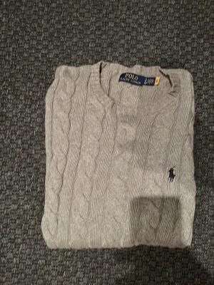 Grå kabelstickad tröja Polo Ralph Lauren - Säljer en klassisk grå kabelstickad tröja från Polo Ralph Lauren. Tröjan har rund hals, ribbade muddar och den ikoniska broderade loggan på bröstet. Perfekt för dig som gillar stilrena och tidlösa plagg.