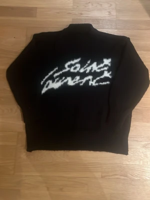 Ysl Svart stickad tröja - Säljer en svart stickad tröja med vit, suddig text över bröstet. Tröjan har lång ärm och en klassisk passform. Perfekt för dig som gillar streetwear och vill ha något unikt i garderoben.
