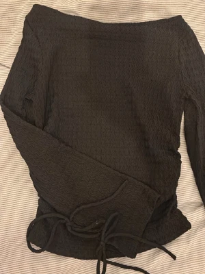 Svart omlottopp med snörning - Säljer en svart topp med lång ärm och snygg snörning i armarna. Tyget har en vågig struktur som ger toppen en cool look. Perfekt att styla med jeans eller kjol för en trendig outfit.