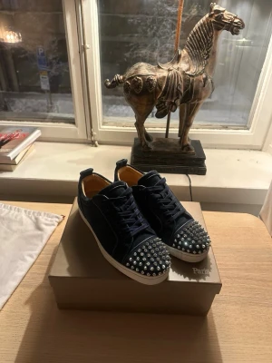 Christian Louboutin sneakers med nitar - Säljer ett par mörkblå sneakers från Christian Louboutin med ikoniska röda sulor och silverfärgade nitar på tån. Skorna har snörning, rund tå och är tillverkade i mocka med skinnfoder. Kommer med originalkartong och dustbag.