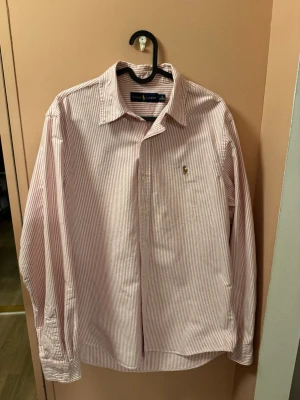 Randig skjorta från Ralph Lauren - Säljer en klassisk skjorta från Ralph Lauren med smala ljusrosa och vita ränder. Skjortan har lång ärm, knappar framtill och broderad logga på bröstet. Tillverkad i bomull och har en tidlös passform som funkar till många stilar. Storlek mellan M och L