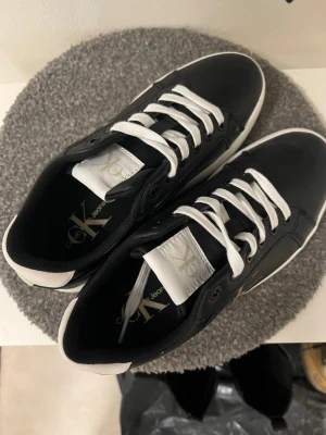 Svarta Calvin Klein sneakers med vita detaljer - Säljer ett par stilrena svarta sneakers från Calvin Klein Jeans med vita snören, vit sula och logga på sidan. Skorna har en vit och mintgrön detalj på hälen och är tillverkade i syntetmaterial. Perfekta för dig som gillar klassisk streetstyle med en twist.