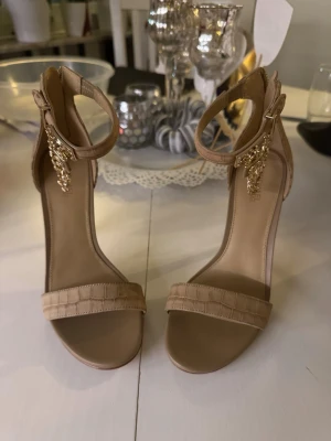 Beige klackskor från Michael Kors - Snygga beige klackskor från Michael Kors med krokopräglat mönster, smal rem över tårna och justerbart ankelband med guldfärgad kedjedetalj och lås. Skorna har hög klack och öppen tå, samt dragkedja bak för enkel påtagning. 6M motsvarar 36 