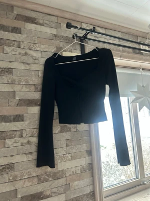 Svart croppad topp med twistad detalj - Säljer en svart croppad topp  lång ärm och twistad detalj framtill. Toppen har en djupare urringning och är gjord i ett stretchigt material som sitter snyggt på kroppen. Perfekt till jeans eller kjol för en trendig look.