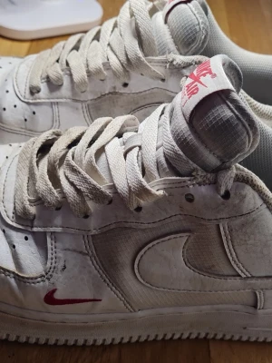 Nike Air Force 1 vita sneakers - Säljer ett par klassiska Nike Air Force 1 sneakers i vitt med röda detaljer. Skorna har snörning, perforerade tår och den ikoniska swoosh-loggan på sidan. Tillverkade i skinn och syntetmaterial, med en platt sula och rund tå. Perfekta för dig som gillar streetwear.